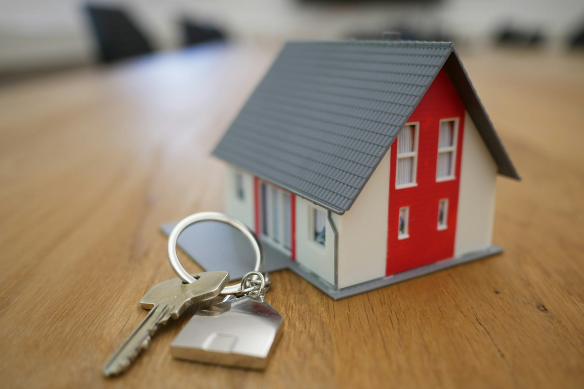A miniature model of a red and white house sits next to a house key on a wood table.